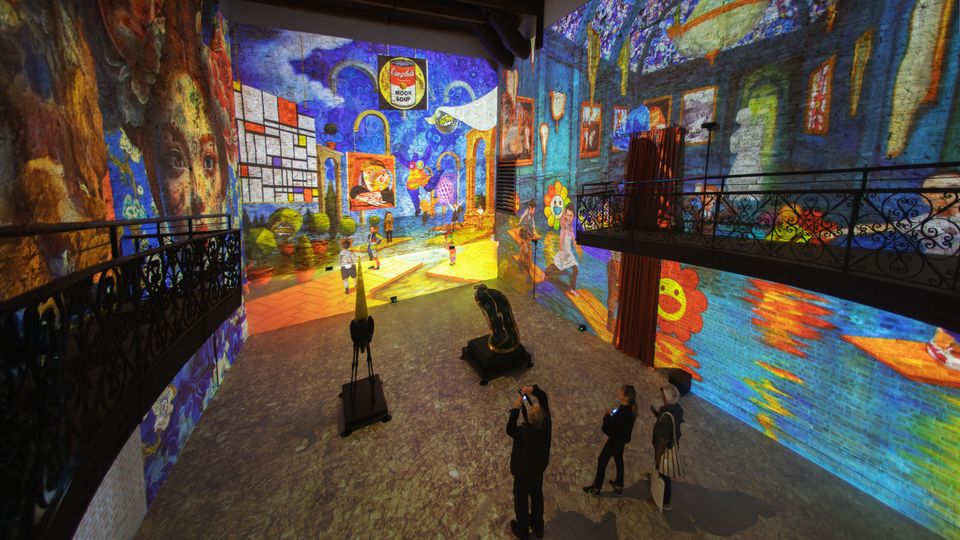 Ampia sala espositiva con soffitto a travi e balconate laterali in ferro battuto, con pavimento in pietra. Pareti interamente coperte da proiezioni artistiche colorate in stile pittorico che trasformano lo spazio in un’installazione immersiva.