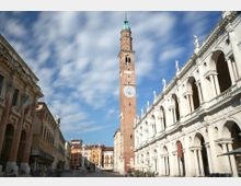 Piazza storica con alta torre in mattoni con orologio e cuspide verde al centro, circondata da edifici rinascimentali. A destra si vede una lunga loggia in pietra bianca con archi sovrapposti e statue sul cornicione, sotto un cielo parzialmente nuvoloso.