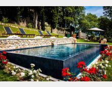 Piscina rettangolare a sfioro con rivestimento scuro, circondata da aiuole fiorite e da un muretto in pietra che separa la vasca da un prato terrazzato con lettini prendisole. Sullo sfondo si vedono alberi alti, un muro in pietra e un’area relax con ombrellone.