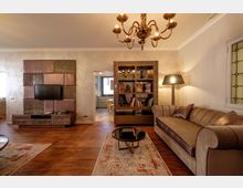 Soggiorno arredato in stile moderno con pavimento in legno, divano in tessuto e pelle, tavolino rotondo su tappeto e lampadario a soffitto. Sulla parete ci sono un mobile TV con pannelli decorativi e una libreria a giorno che separa l’ambiente dall’ingresso verso la cucina.