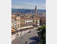 Veduta panoramica di una grande piazza urbana con ampio spazio lastricato e tavolini all’aperto lungo i portici, circondata da edifici storici dai tetti in coppi. Sullo sfondo si vede il centro cittadino con una alta torre campanaria e colline verdi.