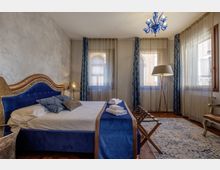 Camera da letto elegante con letto matrimoniale imbottito blu e testiera decorata, biancheria bianca e runner blu, con porta-valigie ai piedi. Tre grandi finestre con tende leggere e drappi blu illuminano l’ambiente, completato da lampada da terra e lampadario in vetro blu sopra un tappeto chiaro.