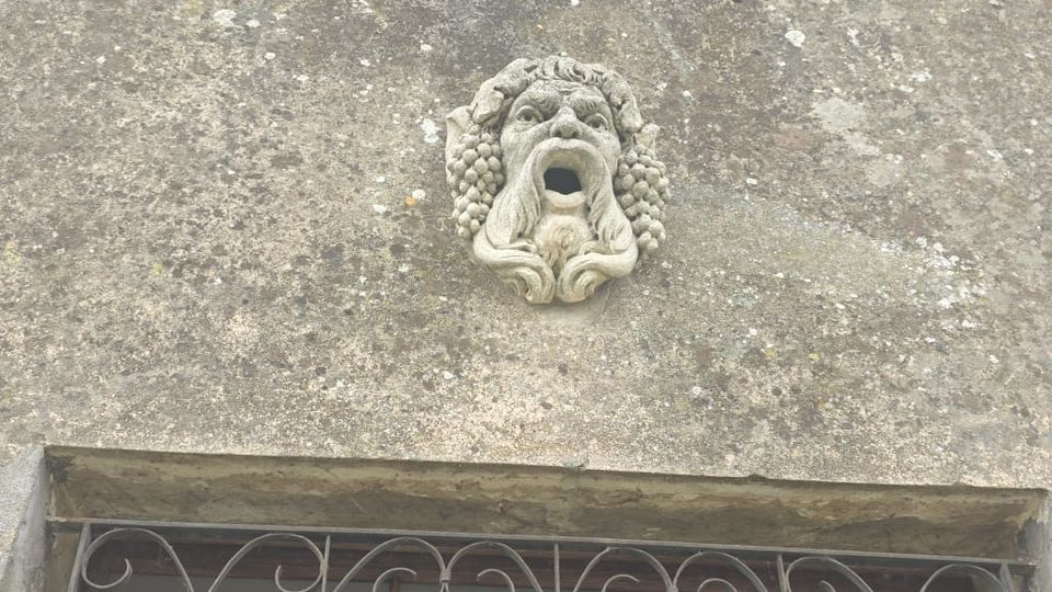 Facciata di un edificio in intonaco invecchiato con una piccola finestra chiusa da persiane azzurre e, sotto, una decorazione in pietra a forma di mascherone. Più in basso si vede un balcone con ringhiera in ferro battuto e parte di un portone in legno con lampadario all’interno.