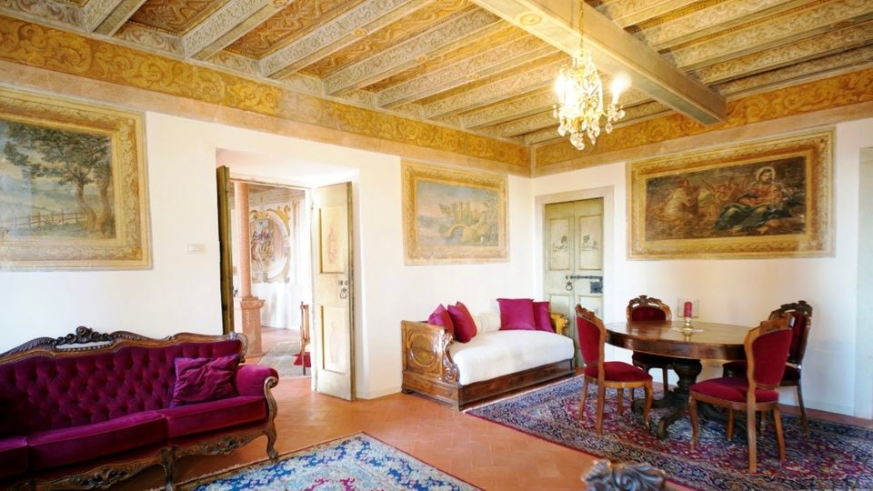 Ampio salone in stile storico con soffitto a travi decorate e lampadario, pareti con grandi affreschi incorniciati e pavimento in cotto con tappeti orientali. Arredi classici in legno con divani e sedie imbottite rosse, tavolo rotondo da pranzo e una porta che conduce a un corridoio adiacente.