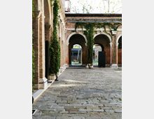 Cortile interno con pavimentazione in pietra e portici in mattoni a vista, scanditi da grandi archi e colonne. Pareti e pilastri sono coperti in parte da edera rampicante, con vista su un edificio sullo sfondo attraverso le arcate.