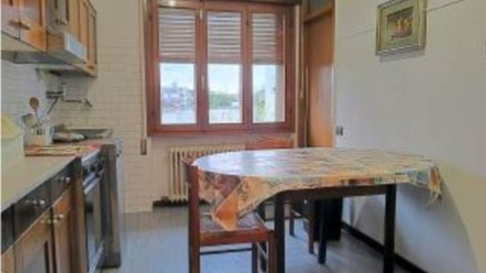 Cucina stretta con mobili in legno e piano cottura sul lato sinistro, pavimento piastrellato e tavolo rettangolare con tovaglia al centro. In fondo una grande finestra con persiane e radiatore sotto, con pareti chiare e un quadro appeso.