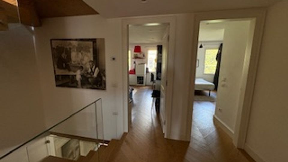 Interno di una casa con corridoio in parquet e pareti bianche, illuminato da luce naturale, con una scala con ringhiera in vetro e legno sulla sinistra. In fondo si aprono porte verso altre stanze, con una camera visibile a destra.
