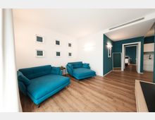 Soggiorno moderno con pavimento in legno, pareti bianche e una parete d’accento color petrolio, arredato con due divani turchesi e piccoli quadri minimalisti. Sul fondo si vede un corridoio con specchio a figura intera che conduce a una camera e a un angolo cucina.