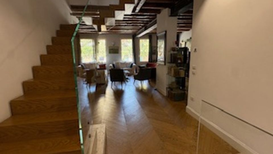 Interno di un’abitazione moderna con scala in legno a vista e pavimento in parquet disposto a spina di pesce. Ambiente open space con soffitto con travi in legno, zona pranzo e ampie vetrate sul fondo che affacciano sul verde esterno.