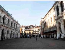 Ampia piazza urbana lastricata, circondata da edifici storici con portici ad archi e facciate in pietra. Ai lati si vedono logge e finestre ad arco, mentre sullo sfondo ci sono palazzi più moderni e attività commerciali al piano terra.