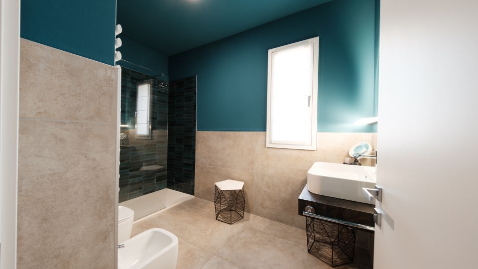 Bagno moderno con pareti color petrolio e rivestimento in piastrelle beige, pavimento in gres e finestra che illumina l’ambiente. A sinistra c’è una doccia con parete in vetro e piastrelle scure, mentre a destra un lavabo rettangolare su mobile con ripiano e sgabelli in metallo.