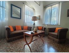 Salotto luminoso con pavimento in graniglia, divano e poltrone marroni con cuscini arancioni disposti attorno a un tavolino in legno. Grandi finestre con tende bianche filtrano la luce; sulla parete ci sono uno specchio e un quadro, con una lampada da terra accanto al divano.