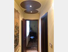 Corridoio interno di un’abitazione con pareti chiare e porte in legno scuro, che conduce a una stanza con pavimento in parquet a spina di pesce. Sul soffitto è presente un controsoffitto decorativo a forma di “8” con faretti incassati.