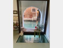 Interno di una stanza con una grande finestra ad arco che si affaccia su un canale veneziano, con un edificio in mattoni visibile all'esterno. La stanza presenta un tavolino verde con oggetti decorativi, un pavimento con un pannello in vetro trasparente e una mensola laterale contenente libri e ornamenti.