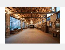 Ampio interno in stile industriale con pareti in mattoni a vista, grandi finestre a griglia su un lato e soffitto alto con travi in legno e capriate. Lungo le pareti si vedono arredi e oggetti d’epoca, con un lampadario centrale e un’area espositiva in fondo al corridoio.