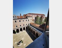 Vista dall’alto di un chiostro in pietra con portici ad archi su due lati e pavimentazione lastricata, con una piccola fontana circolare al centro. Intorno si vedono edifici storici in muratura con tetti in tegole rosse e un albero nel cortile, sotto un cielo sereno.