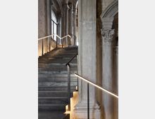 Scala interna in pietra grigia con parapetti in metallo e corrimano illuminati a LED, inserita in un ambiente storico con alti pilastri e colonne ornate. Archi e grandi finestre laterali lasciano filtrare luce naturale lungo la salita.