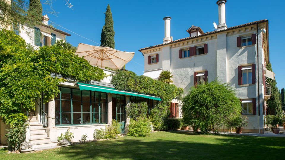 Villa di campagna a più piani con facciata chiara, persiane scure e due camini sul tetto, affacciata su un giardino con prato e alberi. In primo piano una veranda vetrata con colonne e tenda verde, circondata da rampicanti e un grande ombrellone.