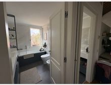 Bagno moderno e luminoso con vasca lungo la parete sinistra, lavandino e WC, piastrelle chiare e pavimento grigio. Una finestra sul fondo illumina la stanza e una porta aperta conduce a una camera da letto adiacente.