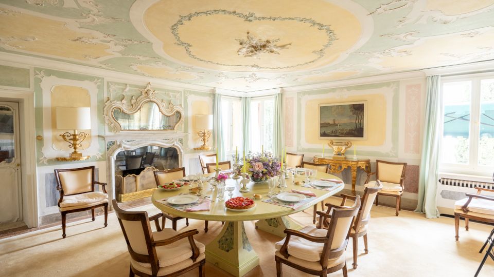 Sala da pranzo in stile classico con soffitto decorato a stucchi e affreschi, camino ornamentale e grande tavolo ovale apparecchiato circondato da sedie imbottite. Ampie finestre con tende chiare illuminano pareti pastello con boiserie e un dipinto, accanto a un mobile consolle dorato.