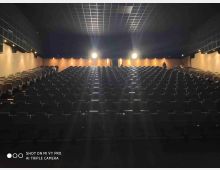 Interno di una grande sala cinematografica o auditorium con file di poltrone scure disposte a gradinata e corridoio centrale. Pareti laterali beige con luci incassate e soffitto alto a griglia, illuminato da due fari in fondo alla sala.