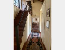 Corridoio interno di una casa con scala in legno e corrimano in metallo a sinistra, grande finestra lungo la scala e soffitto con travi a vista. Sul pavimento in cotto corre un lungo tappeto decorato che conduce a una sala da pranzo visibile in fondo, con porte e armadi bianchi lungo le pareti.