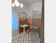 Bagno interno con lavabo su piedistallo e specchio con cornice decorata, pareti rivestite a metà in piastrelle marmorizzate color terracotta e parte superiore bianca. Pavimento a motivi geometrici con stelle e, a destra, una parete divisoria in vetro satinato rigato vicino alla zona vasca o doccia.