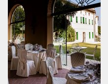 Sala da pranzo con grandi archi e vetrate affacciate su un giardino, con tavoli rotondi apparecchiati e sedie rivestite di bianco. All’esterno si vedono arredi da patio in ferro battuto e un edificio a due piani con persiane verdi.