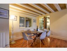 Sala da pranzo interna con pavimento in legno e soffitto con travi a vista dipinte di chiaro, con tavolo ovale apparecchiato e sei sedie moderne. Sulla parete ci sono quadri, una grande lampada da terra, una finestra interna con pannello decorativo e una vetrina illuminata con ripiani in vetro e colonna in legno.