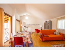 Soggiorno open space mansardato con pavimento in legno, tavolo da pranzo apparecchiato vicino a grandi porte finestre che danno su un balcone e un’area relax con divano arancione e poltrona rossa. Sul fondo si vedono una libreria/vetrina grigia, lampade da terra e una finestra con tende leggere.