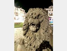 Primo piano di una scultura in pietra raffigurante un leone con criniera scolpita e una sfera in bocca, collocata su una pavimentazione in pietra in una piazza illuminata di notte. Sullo sfondo si vedono edifici storici e un’area verde con una fontana.