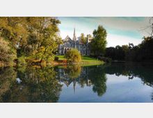 Edificio storico in stile neogotico con una guglia centrale, visto da un prato alberato sulla riva di un lago. L’acqua calma riflette la facciata e gli alberi circostanti in un parco paesaggistico.