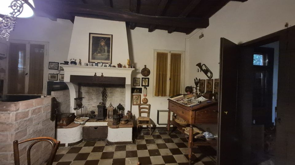 Interno rustico con soffitto a travi in legno e pavimento a scacchi bianco e nero, dominato da un grande camino in muratura con mensola decorata. Sulla destra ci sono una finestra con tende e un tavolo da lavoro inclinato con oggetti, mentre una porta scura aperta conduce a un’altra stanza.