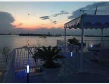 Terrazza sul lungomare con ringhiera bianca, pergola con tenda e tavolini, decorata con piante in vaso e piccole luci blu lungo il parapetto. Sullo sfondo si vede una distesa d’acqua al tramonto con una barca illuminata e la skyline della città in lontananza.