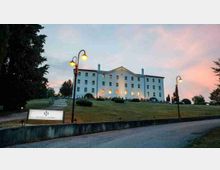 Grande edificio alberghiero bianco a più piani su una collina erbosa, con facciata simmetrica e finestre illuminate al crepuscolo. In primo piano si vedono strada e muretto, lampioni accesi e un’insegna dell’hotel lungo il vialetto d’accesso.