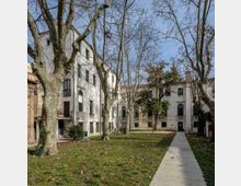 Cortile interno con prato e grandi alberi spogli, attraversato da un vialetto rettilineo in cemento che conduce a edifici chiari a più piani con finestre ad arco e piccoli balconi. Sullo sfondo si vedono facciate intonacate e tetti in coppi sotto un cielo sereno.
