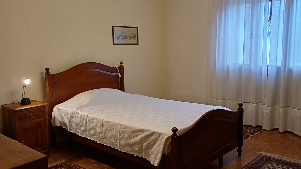 Camera da letto con letto matrimoniale in legno e copriletto chiaro, comodino con lampada e due tappeti decorati su pavimento in parquet. Una grande finestra con tende bianche illumina la stanza, con lampadario a soffitto e pareti chiare.