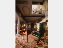 Interno di un salone in stile storico con colonne in pietra, soffitti decorati e travi a vista, organizzato attorno a un grande tavolo in legno su un tappeto. Una balconata con ringhiera metallica si affaccia dall’alto, con pavimento a scacchi e arredi classici come una poltrona in pelle verde.