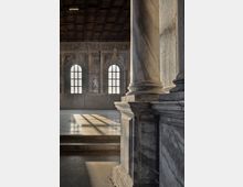 Interno di una grande sala storica con soffitto a travi in legno e pareti affrescate, illuminate da alte finestre ad arco. In primo piano si vede un massiccio pilastro in pietra e il pavimento in marmo con fasci di luce e ombre proiettate dalle finestre.