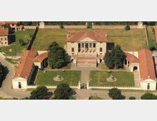 Vista aerea di una villa storica con corpo centrale a frontone e portico a colonne, affiancata da due ali porticate con tetti in coppi rossi. Davanti si estende un giardino rettangolare con vialetti, fontane e alberi, circondato da recinzioni e muri perimetrali.