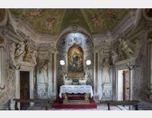 Interno di una piccola cappella barocca con altare centrale su pedana, tovaglia ricamata e pala d’altare incorniciata da un arco. Pareti e soffitto sono riccamente decorati con stucchi e affreschi, statue ai lati e due piccole finestre ottagonali che illuminano l’ambiente.