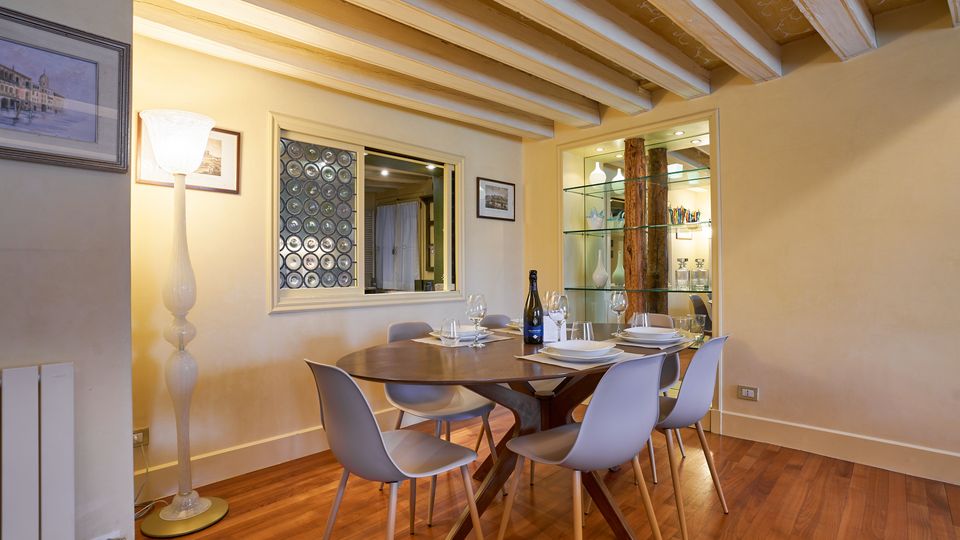 Sala da pranzo interna con pavimento in legno e soffitto con travi a vista dipinte di chiaro, con tavolo ovale apparecchiato e sei sedie moderne. Sulla parete ci sono quadri, una grande lampada da terra, una finestra interna con pannello decorativo e una vetrina illuminata con ripiani in vetro e colonna in legno.