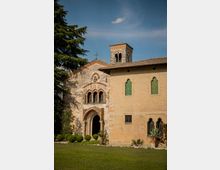 Facciata di una chiesa in pietra e laterizio con portale ad arco, piccole arcate decorative e campanile quadrato con bifore, affiancata da un edificio adiacente con finestre ad arco e persiane verdi. Davanti si vede un prato con siepi e alberi, sotto un cielo sereno.