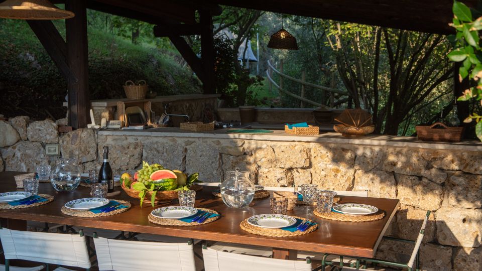 Zona pranzo all’aperto sotto una tettoia in legno, con grande tavolo apparecchiato e sedie bianche. Sullo sfondo c’è una cucina in muratura con piano in pietra, lampade intrecciate a sospensione e vegetazione alberata tutt’intorno.