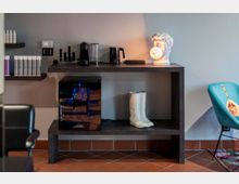 Angolo interno moderno con mensola e scaffali a parete: sul ripiano ci sono una macchina da caffè, bollitore e lampada a forma di busto classico, con un piccolo frigo per vino e una scultura di stivali bianchi nel vano inferiore. Pavimento in piastrelle color terracotta e una poltroncina turchese visibile sul lato destro.