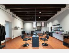 Interno di un salone di parrucchiere moderno con travi a vista in legno scuro, pavimento in cotto e pareti grigie. Al centro ci sono postazioni con sedie e specchi su un bancone, mentre lungo le pareti si vedono mensole con prodotti e due finestre sul fondo.