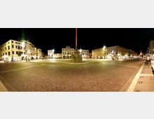 Ampia piazza cittadina pavimentata in ciottoli, illuminata da lampioni notturni, con un alto monumento su basamento al centro. Intorno si affacciano edifici storici a più piani con portici e arcate lungo il perimetro della piazza.