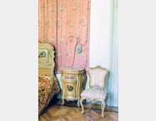Interno di una camera in stile vintage con pavimento in parquet, letto e comodino decorati con motivi floreali e una sedia imbottita a righe. Sullo sfondo si vede una tenda rosa a fiori e una porta bianca sulla destra.