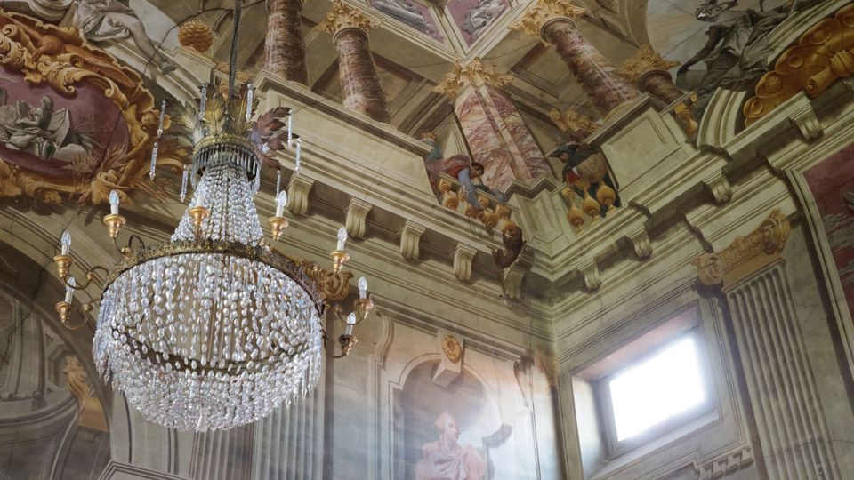 Interno di una chiesa barocca con soffitto affrescato raffigurante colonne e scene allegoriche, decorazioni dorate e un grande lampadario di cristallo sospeso al centro. Un'apertura rettangolare lascia entrare la luce naturale sulla parete laterale.