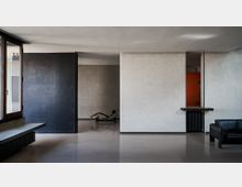 Interno di un appartamento moderno e minimalista con pavimento lucido, grandi pareti divisorie in intonaco grigio e bianco e una ampia finestra laterale. Nello spazio si vedono una chaise longue al centro, un piccolo bancone con termosifone a destra e un divano in pelle scura in primo piano.